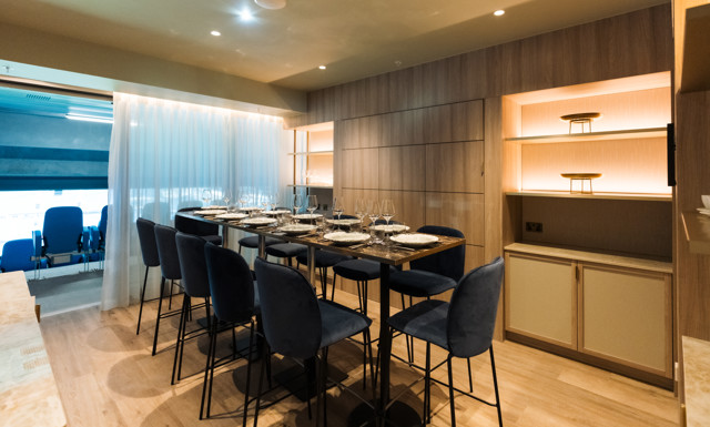 Elegant dining area with high table, set with glassware and plates, surrounded by upholstered chairs. Soft lighting and wood paneling, with floor-to-ceiling windows overlooking stadium seats.