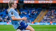 Football player in light blue jersey slides on knees in celebration at a stadium with Etihad branding visible.