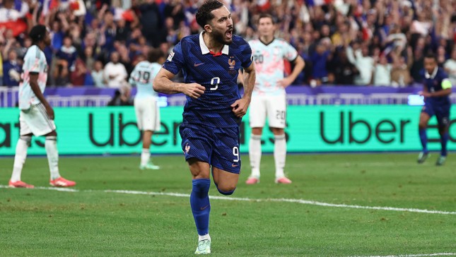 A soccer player in a blue jersey with number 9 celebrates after scoring a goal on the field. Opponents in light-colored jerseys appear disappointed in the background.