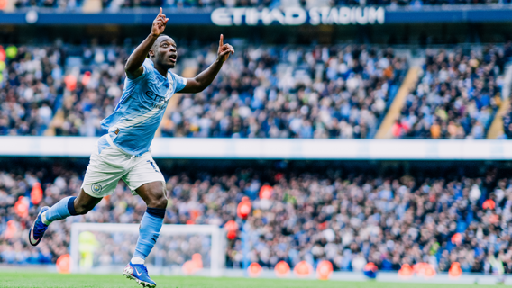 A football player in a blue jersey is celebrating a goal at a packed Etihad Stadium with raised arms and pointing upwards.