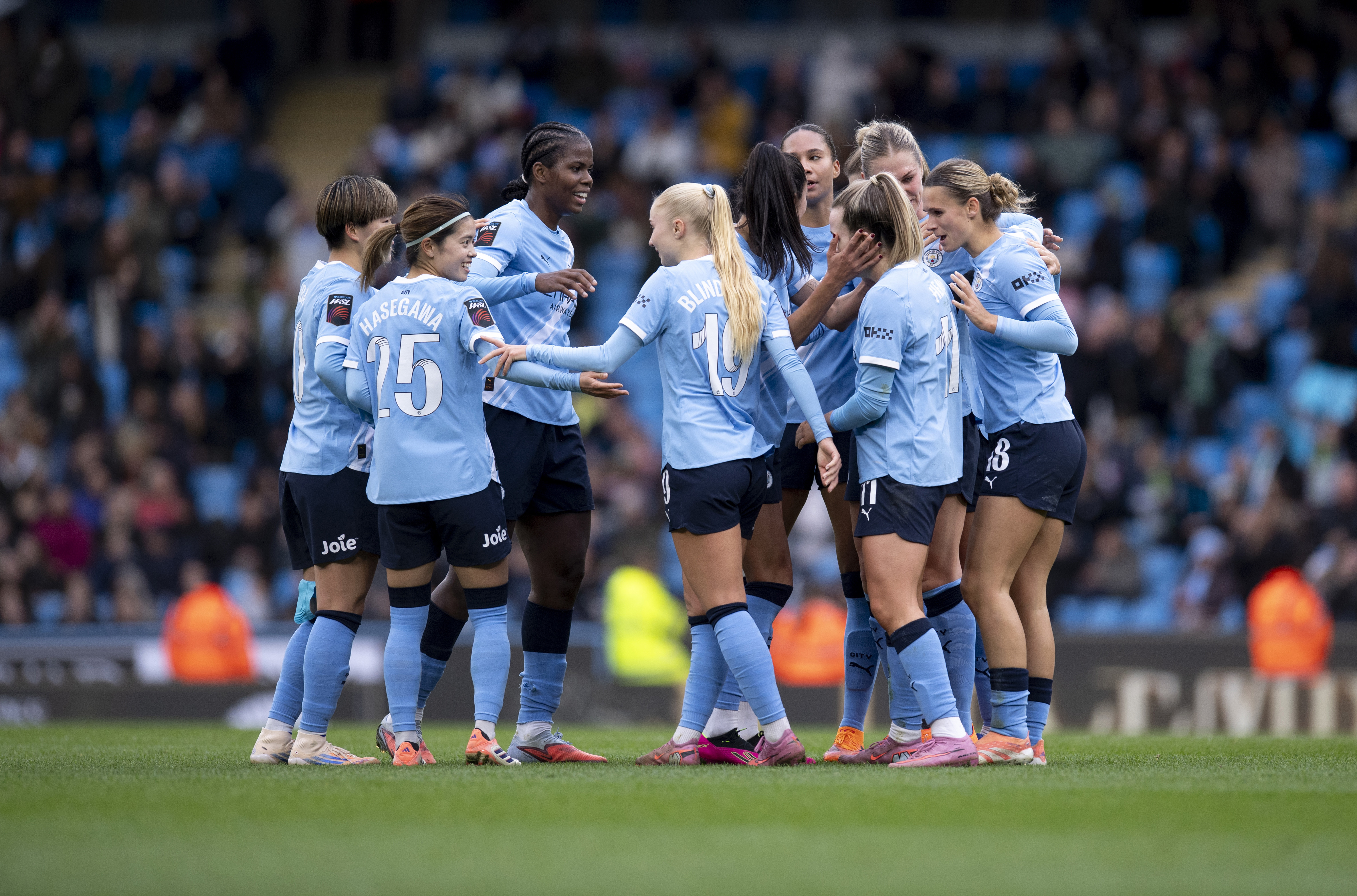 Manchester City women's team players huddle together on the field wearing light blue jerseys during a match at a stadium.