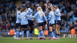 Manchester City women's team players huddle together on the field wearing light blue jerseys during a match at a stadium.