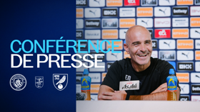 A Manchester City press conference with a smiling man speaking, bottles of Lucozade on the table, and logos of Manchester City, Carabao Cup, and Norwich City displayed.