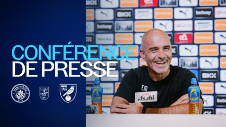 A Manchester City press conference with a smiling man speaking, bottles of Lucozade on the table, and logos of Manchester City, Carabao Cup, and Norwich City displayed.