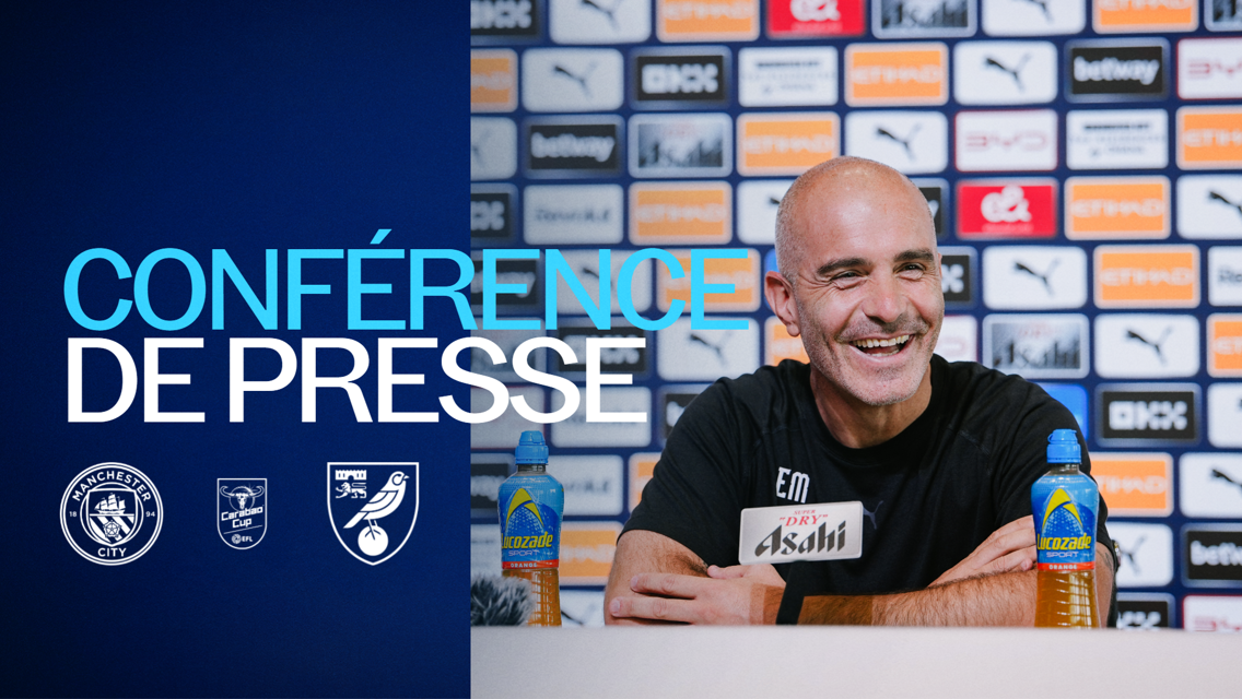 A Manchester City press conference with a smiling man speaking, bottles of Lucozade on the table, and logos of Manchester City, Carabao Cup, and Norwich City displayed.