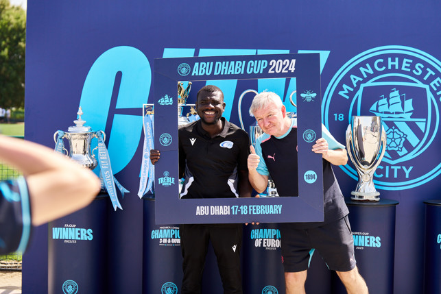 Two people holding an Abu Dhabi Cup 2024 frame, standing in front of Manchester City trophies, promoting the event happening in Abu Dhabi on February 17-18.
