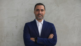A businessman wearing a navy blue suit and a white shirt stands confidently with arms crossed against a textured gray wall.