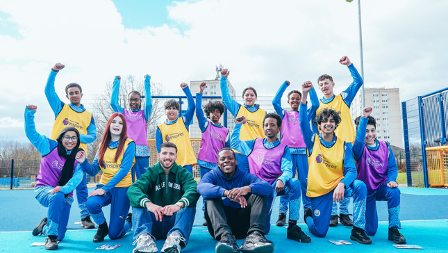 A group of young people in sports outfits with 'inspires' bibs pose with raised fists outdoors on a blue sports field.