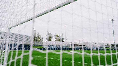 View through a football goal net showing a soccer field with surrounding structures and a pile of footballs, associated with Manchester City facilities.