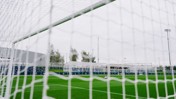 View through a football goal net showing a soccer field with surrounding structures and a pile of footballs, associated with Manchester City facilities.