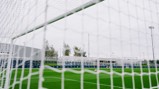 View through a football goal net showing a soccer field with surrounding structures and a pile of footballs, associated with Manchester City facilities.