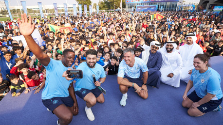 A large group of people, including individuals in blue sports attire, pose for a photo at an outdoor event with a crowd in the background.