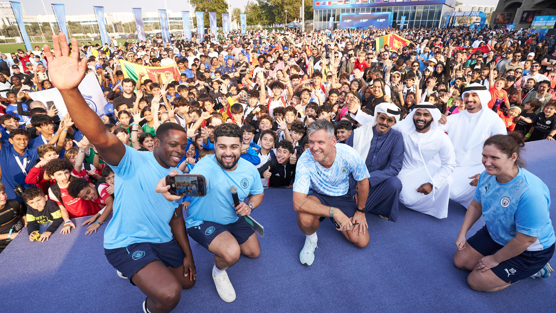 A large group of people, including individuals in blue sports attire, pose for a photo at an outdoor event with a crowd in the background.