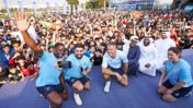 A large group of people, including individuals in blue sports attire, pose for a photo at an outdoor event with a crowd in the background.