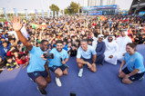 A large group of people, including individuals in blue sports attire, pose for a photo at an outdoor event with a crowd in the background.