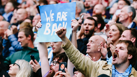 A crowd of enthusiastic fans, with one holding up a sign that reads 'WE ARE BACK'.