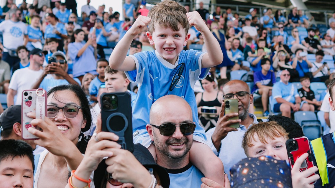Young fan in Manchester City jersey raised on shoulders amid cheering crowd, capturing celebration and excitement at sports event.