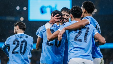 Manchester City players celebrate a goal on the field, wearing light blue jerseys. One player's face is blurred.