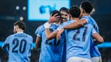 Manchester City players celebrate a goal on the field, wearing light blue jerseys. One player's face is blurred.
