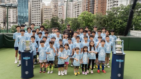 A group of children in Manchester City shirts posing on a football field with two trophies. An adult in black stands in the middle, with urban buildings in the background.