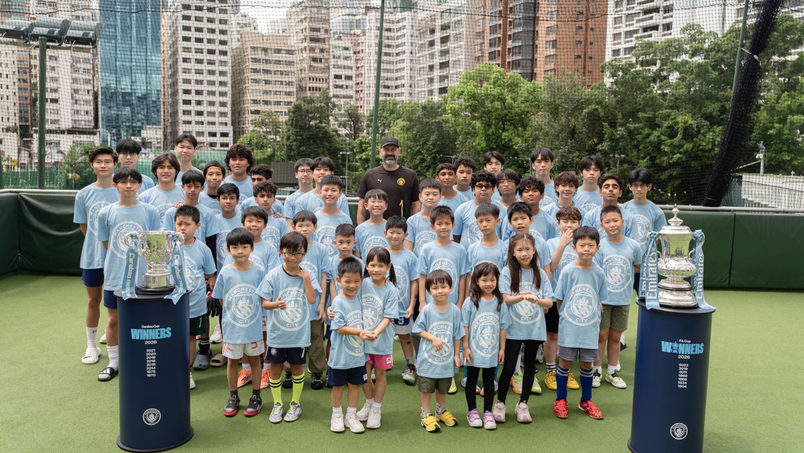 A group of children in Manchester City shirts posing on a football field with two trophies. An adult in black stands in the middle, with urban buildings in the background.