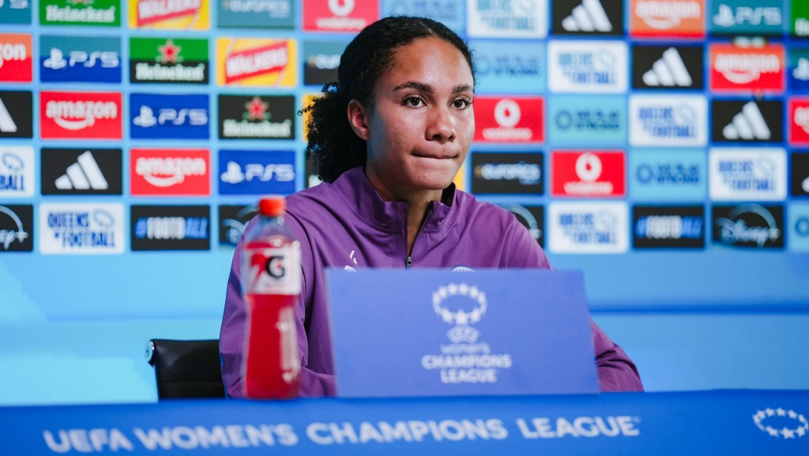 A female football player at a UEFA Women's Champions League press conference, sitting in front of a sponsor backdrop with a Gatorade bottle on the table.