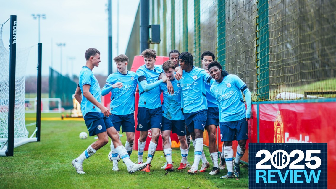 A group of young soccer players in Manchester City kits celebrating near a goalpost on a grass field. The players are wearing blue jerseys and shorts with Manchester City and sponsor logos.