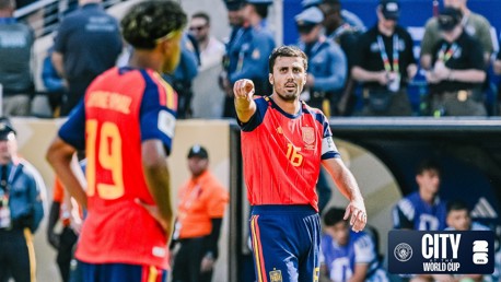 Two football players in red and blue jerseys at a World Cup event. One player points towards the field. City World Cup logo in the corner.