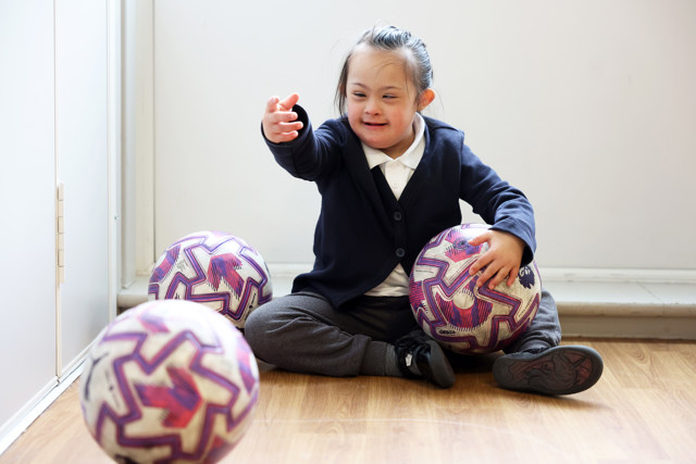 A child sitting on the floor with three purple and white patterned sports balls, reaching out with one hand.
