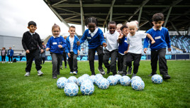A group of children, all dressed in blue and white uniforms, are playing with multiple soccer balls on a field.