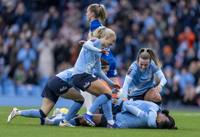 A group of football players on the field, with one player lying on the ground and others assisting. They are wearing blue uniforms and playing on a grass pitch with a crowd in the background.