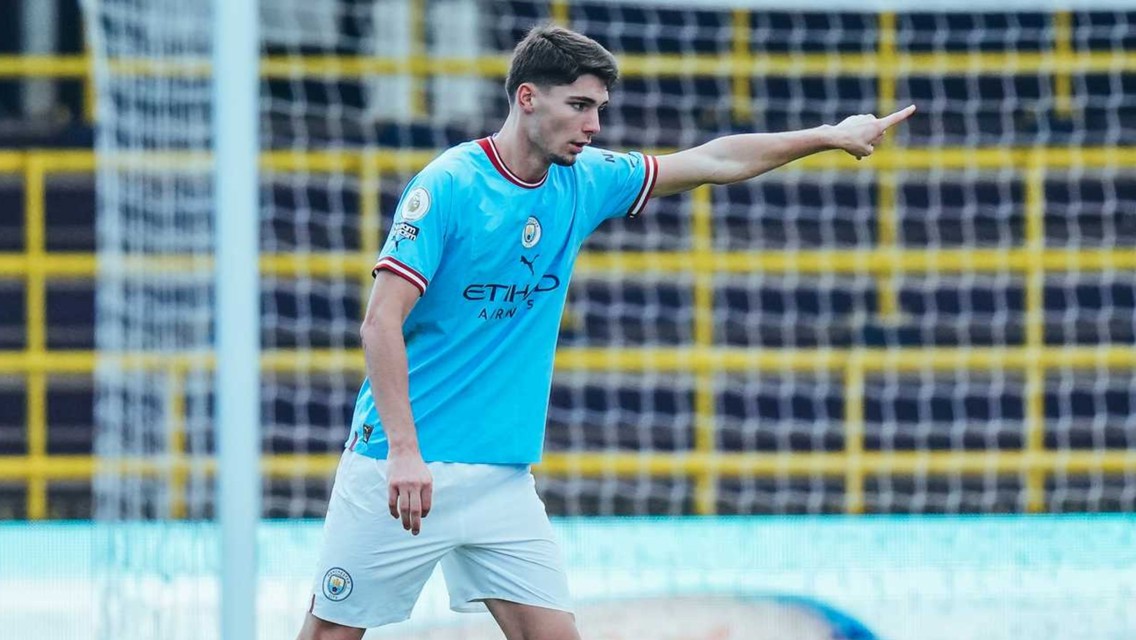 A soccer player in a Manchester City kit gestures during a match on the field, with a net and stands in the background.