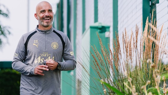 A coach in Manchester City gear with a background of green fences and tall grass.