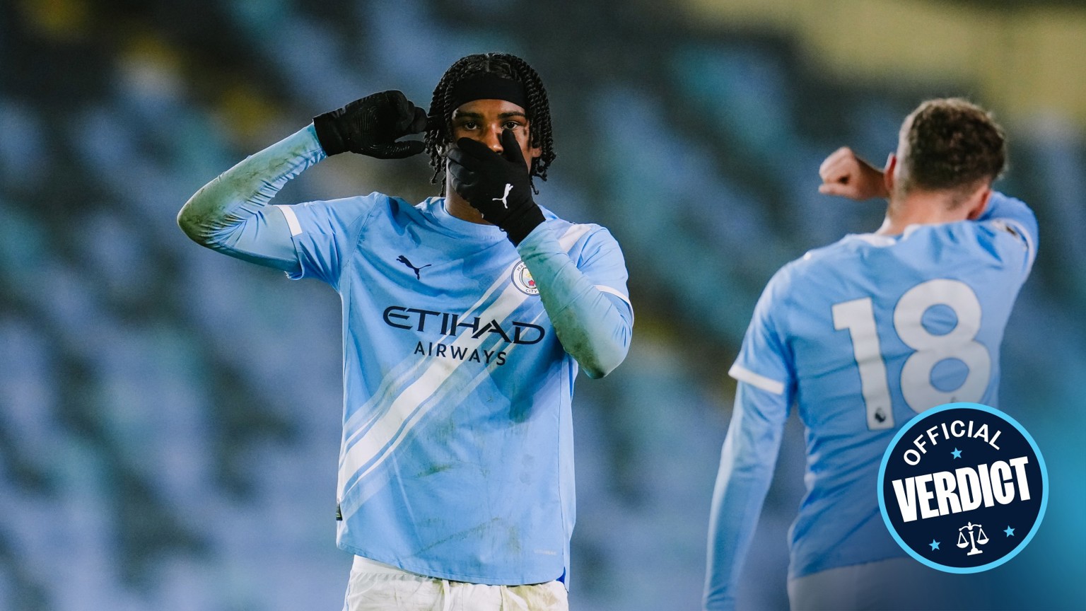 Image shows Manchester City football players celebrating a goal. The player in the foreground wears Manchester City kit and mimics headphones with his hands. Manchester City logo is visible. Another player with jersey number 18 is in the background.
