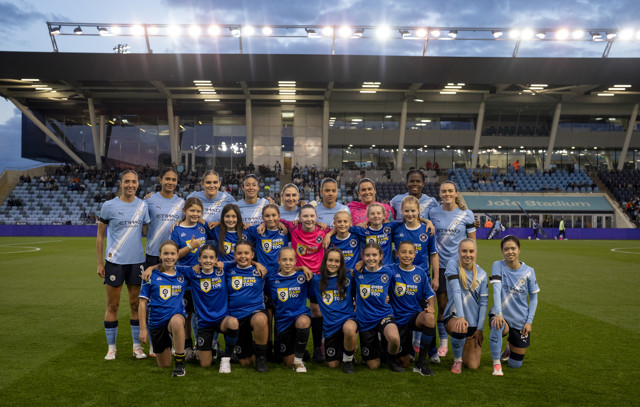 Manchester City team photo with children in blue jerseys at Joie Stadium, taken before a match. The players' faces are blurred.
