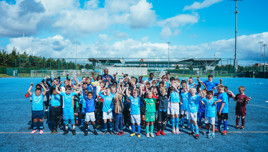 A large group of children wearing soccer uniforms posing on a blue soccer field. The kids are excited, with some raising their arms. The field has a stadium in the background under a blue sky with clouds.