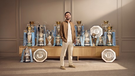 A person standing in front of a display of multiple football trophies, including Premier League, FA Cup, and Community Shield trophies, all adorned with Manchester City branding.