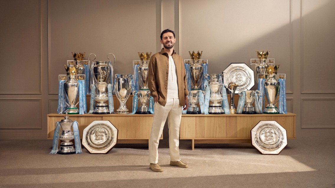 A person standing in front of a display of multiple football trophies, including Premier League, FA Cup, and Community Shield trophies, all adorned with Manchester City branding.