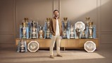 A person standing in front of a display of multiple football trophies, including Premier League, FA Cup, and Community Shield trophies, all adorned with Manchester City branding.