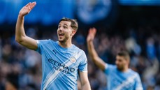 Two football players in Manchester City uniforms are on the field, waving to the audience.