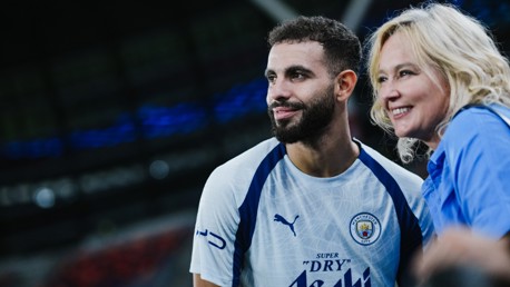 A male football player wearing a Manchester City jersey poses with a female fan at a stadium.
