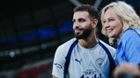 A male football player wearing a Manchester City jersey poses with a female fan at a stadium.