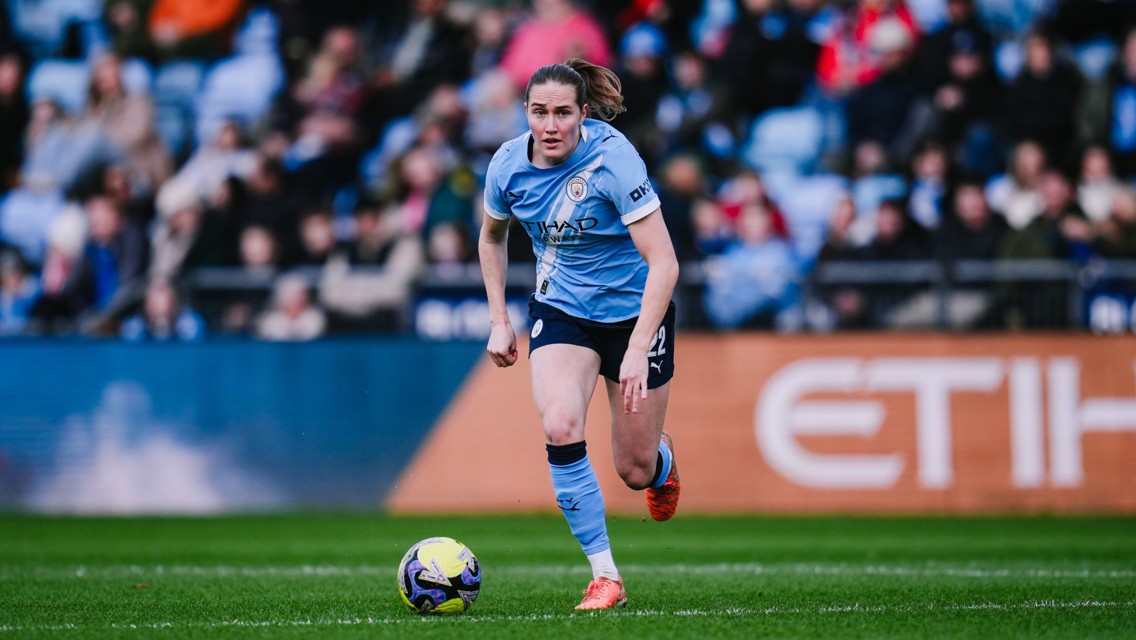 A football player from Manchester City Women in action during a match, wearing a light blue kit and running towards the ball on a field with spectators in the background.