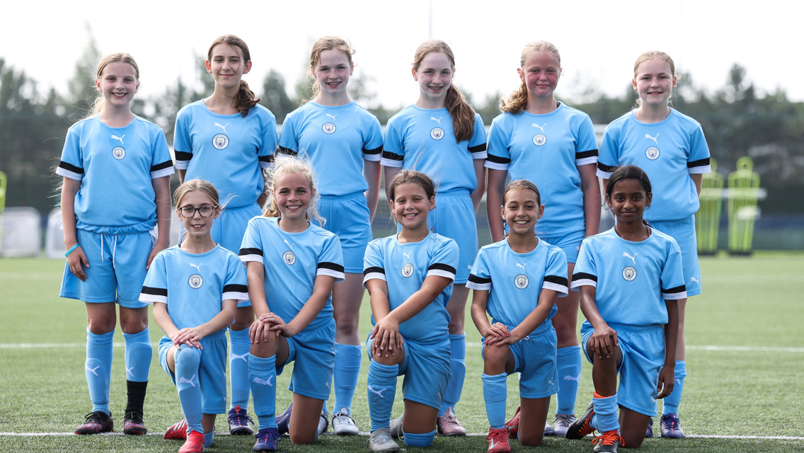 A group of young soccer players wearing blue uniforms with Puma logos and a Manchester City crest, posing for a team photo on a field.