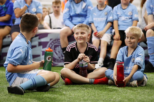 Three young boys sitting on the grass, drinking from sports bottles, surrounded by other children wearing Manchester City jerseys.