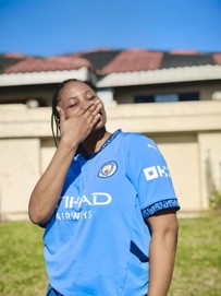 A person is outdoors wearing a Manchester City football shirt with visible logos and sponsors. The background includes a house and blue sky.