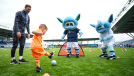 A young child kicks a soccer ball on a field, guided by a coach and two Manchester City mascots during an event at the City Football Academy.