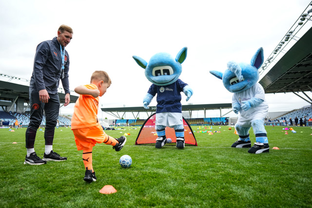 A young child kicks a soccer ball on a field, guided by a coach and two Manchester City mascots during an event at the City Football Academy.