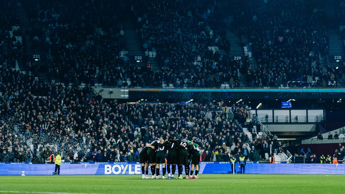A soccer team huddles on the field in a stadium filled with spectators, preparing for the match.