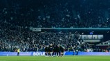 A soccer team huddles on the field in a stadium filled with spectators, preparing for the match.